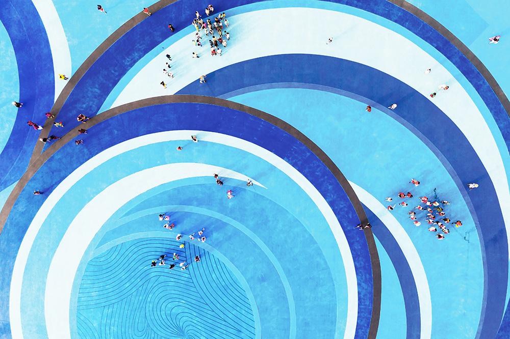 A blue swimming pool filled with people enjoying a sunny day, some swimming and others relaxing at the edge.